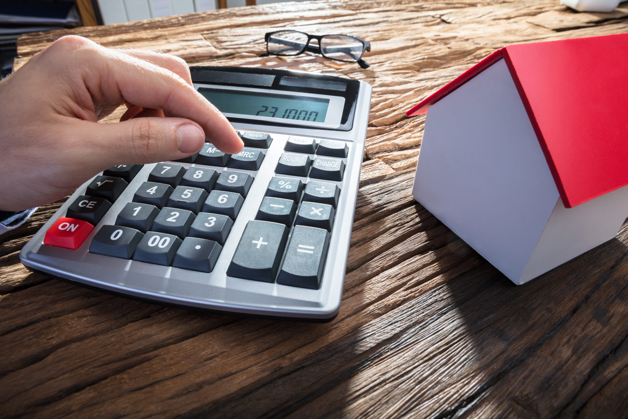 Persons Hand Using Calculator With House Model On Wooden Desk